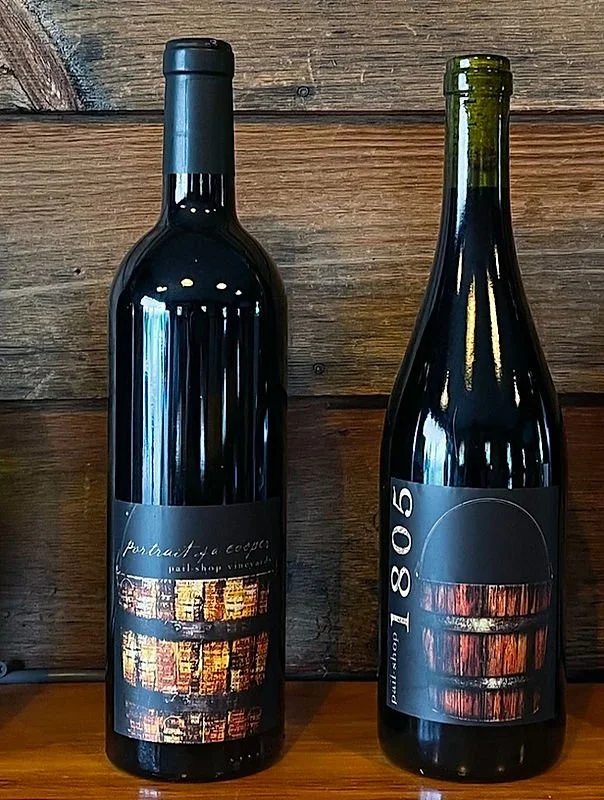 Two bottles of wine with artistic labels, placed on a wooden shelf against a wooden wall.