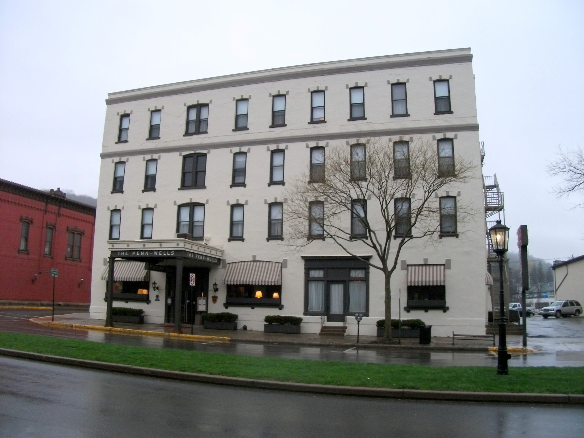 A white multi-story building with black window frames, an awning at the entrance reading ''The Penn-Wells,'' a leafless tree in front, and a street with parked cars and a lamppost, under overcast sky.