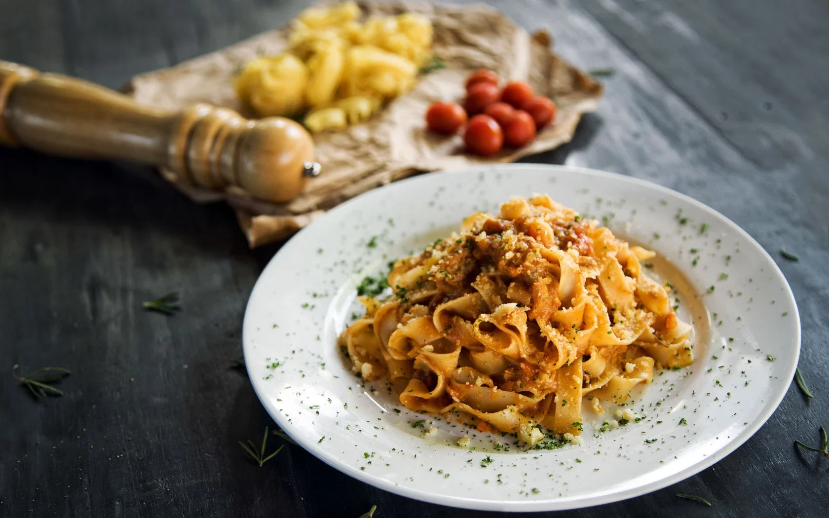 A plate of cooked pasta with meat sauce garnished with herbs on a white dish. In the background, a wooden pepper mill, cherry tomatoes, and pasta are on a rustic wooden surface.