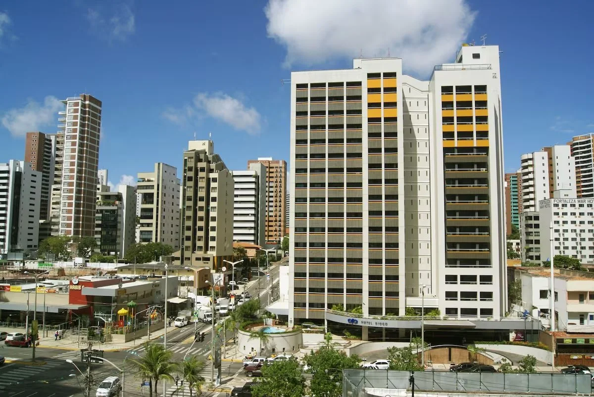 Cityscape of tall modern buildings with a hotel in the foreground, busy streets with cars, palm trees, and a partly cloudy sky.