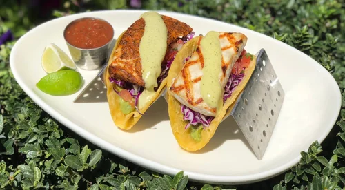 Two fish tacos with shredded purple cabbage, diced tomatoes, and green sauce, served with lime wedges and a side of salsa on a white dish.
