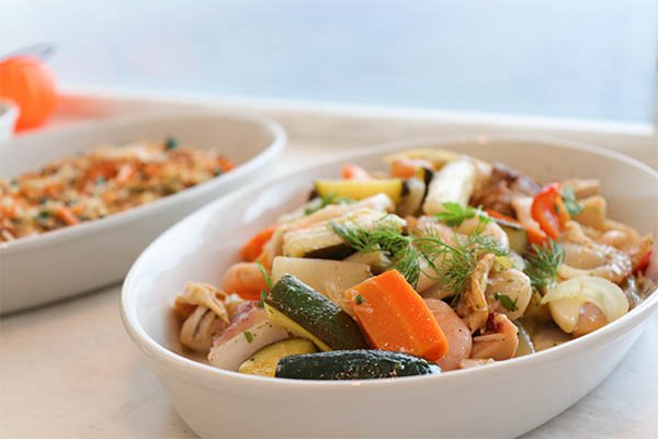 A white oval bowl filled with cooked mixed vegetables including zucchini, carrots, and onions, garnished with fresh herbs, next to a white dish containing rice and vegetables in the background.
