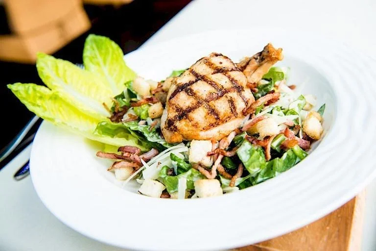 Grilled chicken Caesar salad with romaine lettuce, bacon bits, croutons, and grilled chicken on top, served in a white bowl.