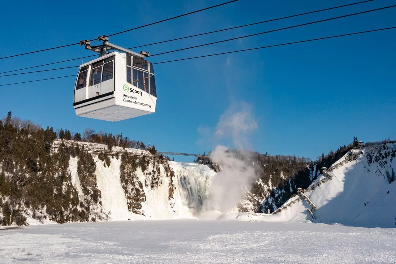 Cable car in winter over frozen water with snowy cliffs and trees, steam or mist near a waterfall in the background, clear blue sky.