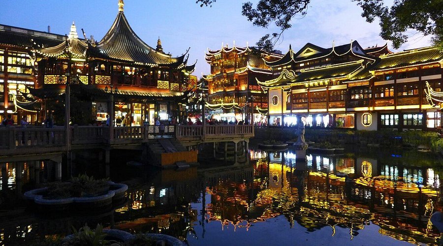 Nighttime view of illuminated traditional Chinese-style buildings with ornate roofs, surrounding a pond that reflects their lights, in an outdoor setting.