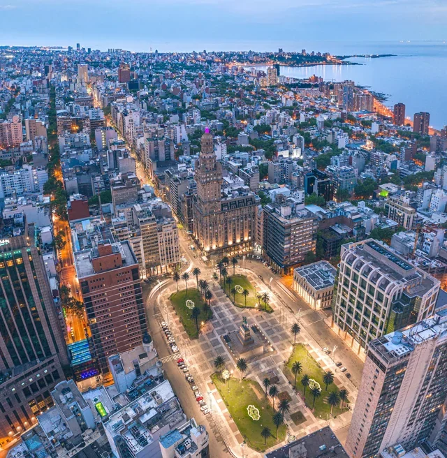 Aerial view of a cityscape showing a city park with palm trees and walkways, surrounded by tall buildings along a city street, with water in the background at dusk.