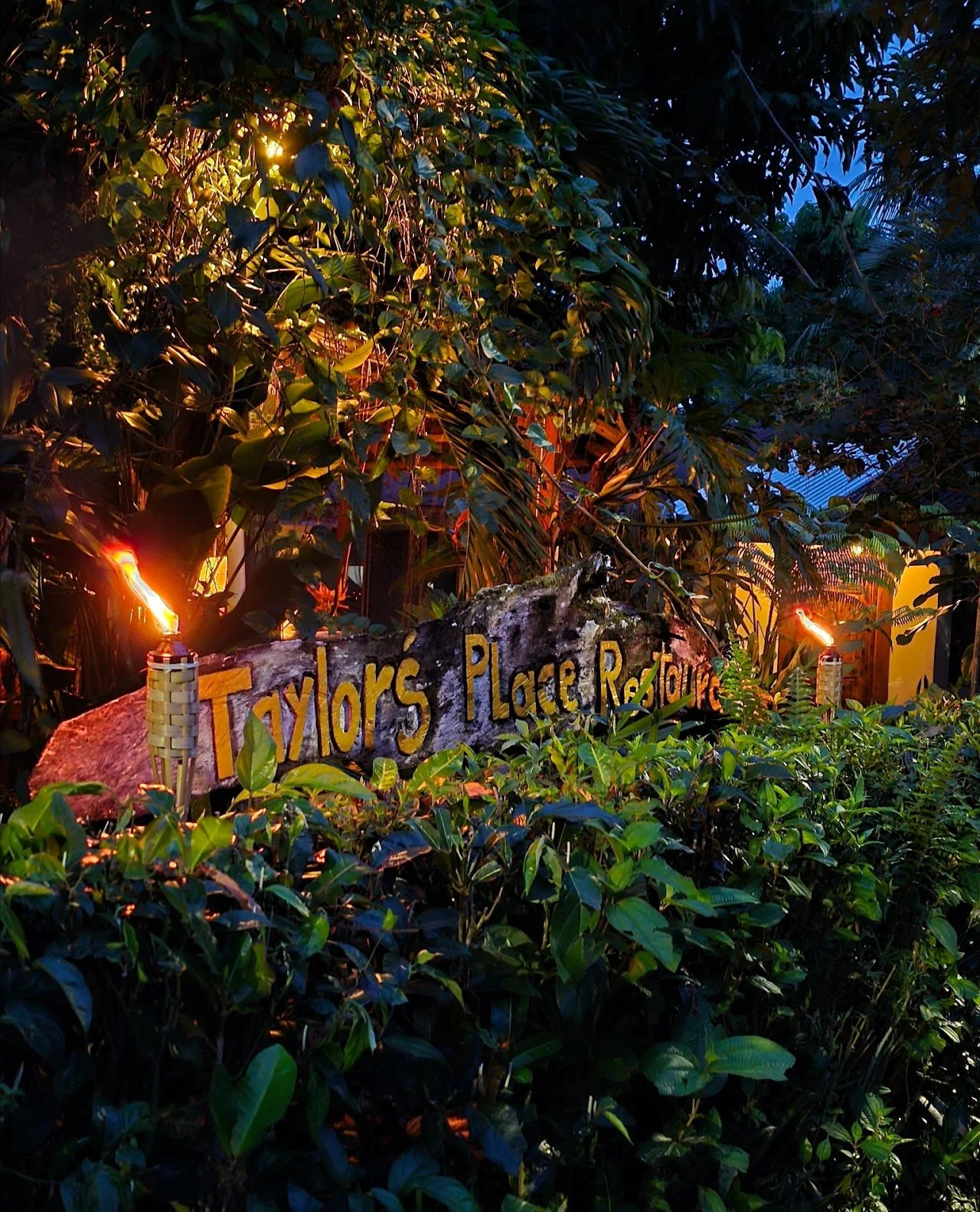 A wooden sign reading 'Taylors Place Retreat' in colorful letters, surrounded by lush green plants and trees, with tiki torches providing warm lighting in a garden setting during dusk.