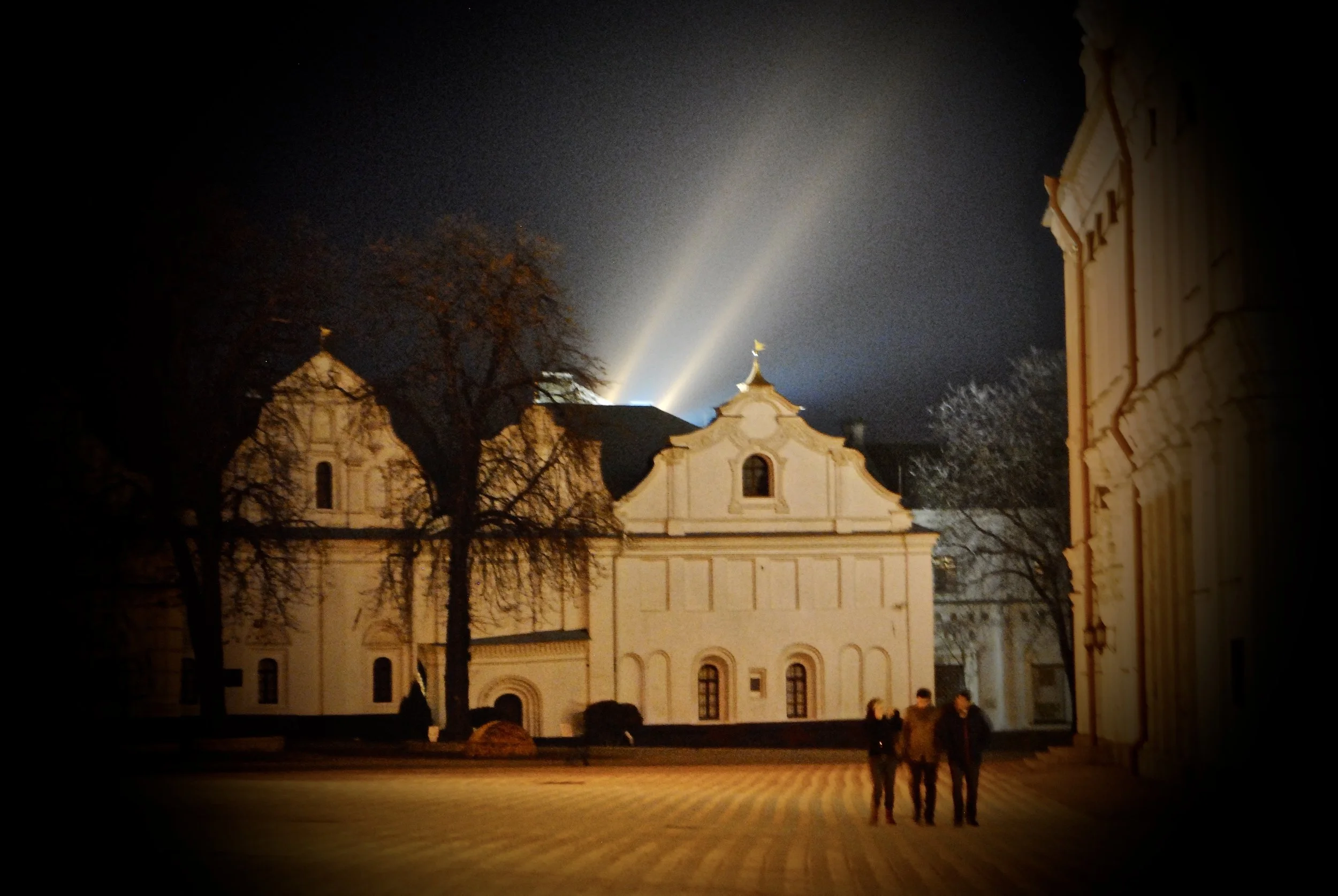 Night view of a historic building illuminated by outdoor lights, with trees in the foreground and three people walking in front.