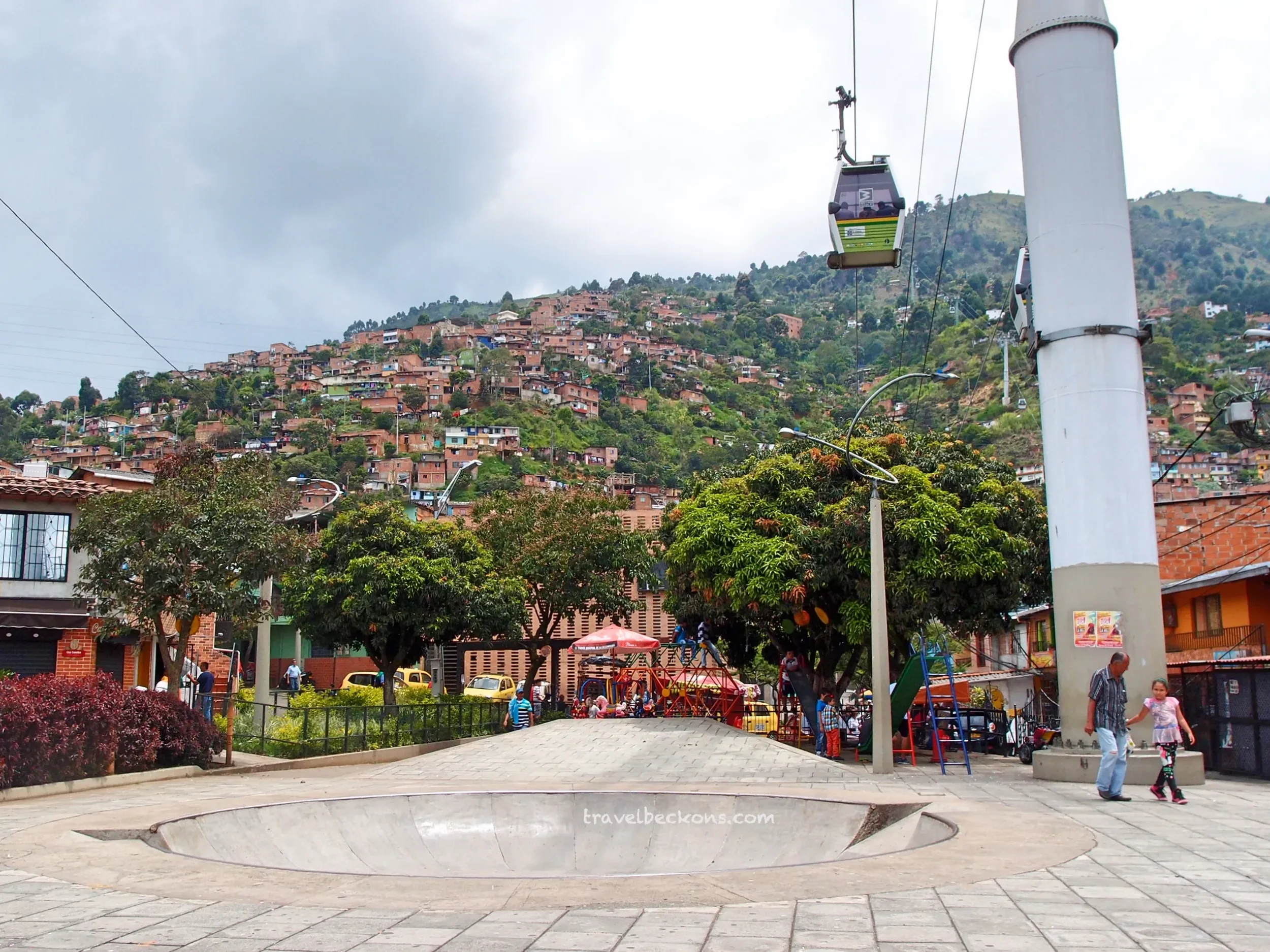 A small amusement park or playground with a carousel, children playing, and people walking. A hillside covered with houses in the background.