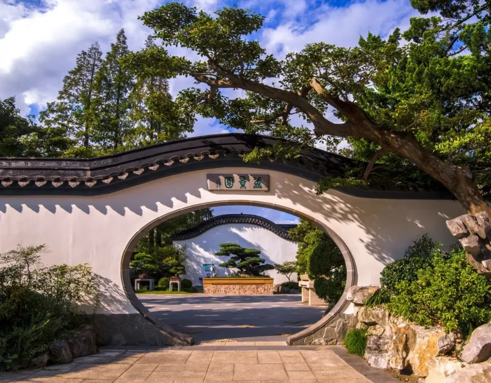 Traditional Chinese garden entrance with an arched white wall, decorative roof edge, Chinese characters, lush greenery, and a pathway inside.
