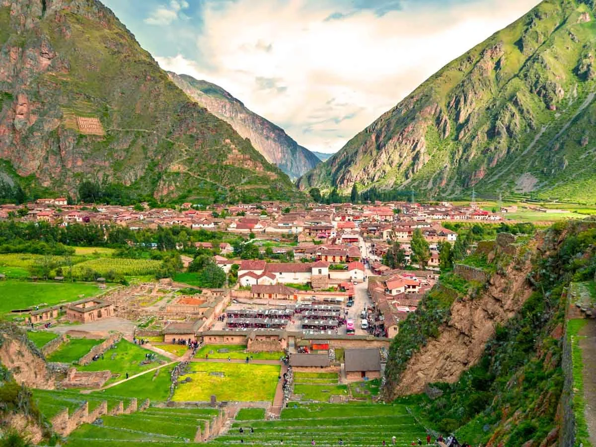 A panoramic view of a lush green valley with a small town nestled between tall, rocky mountains under a partly cloudy sky.