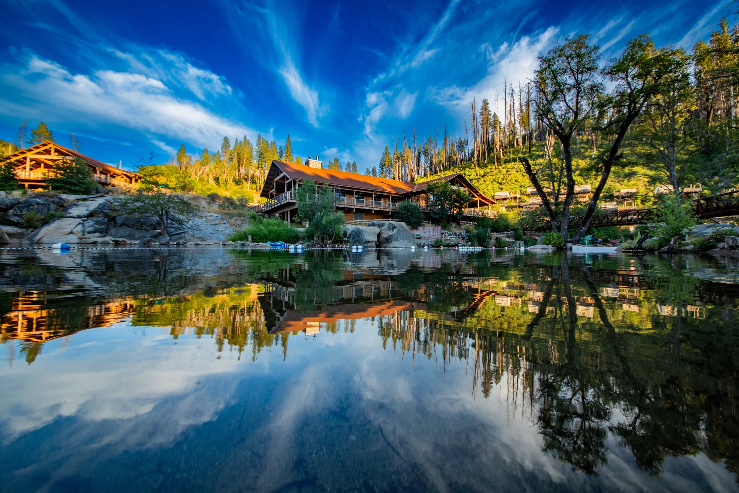 Scenic view of a mountain lodge by a reflective lake, surrounded by pine trees under a partly cloudy sky.
