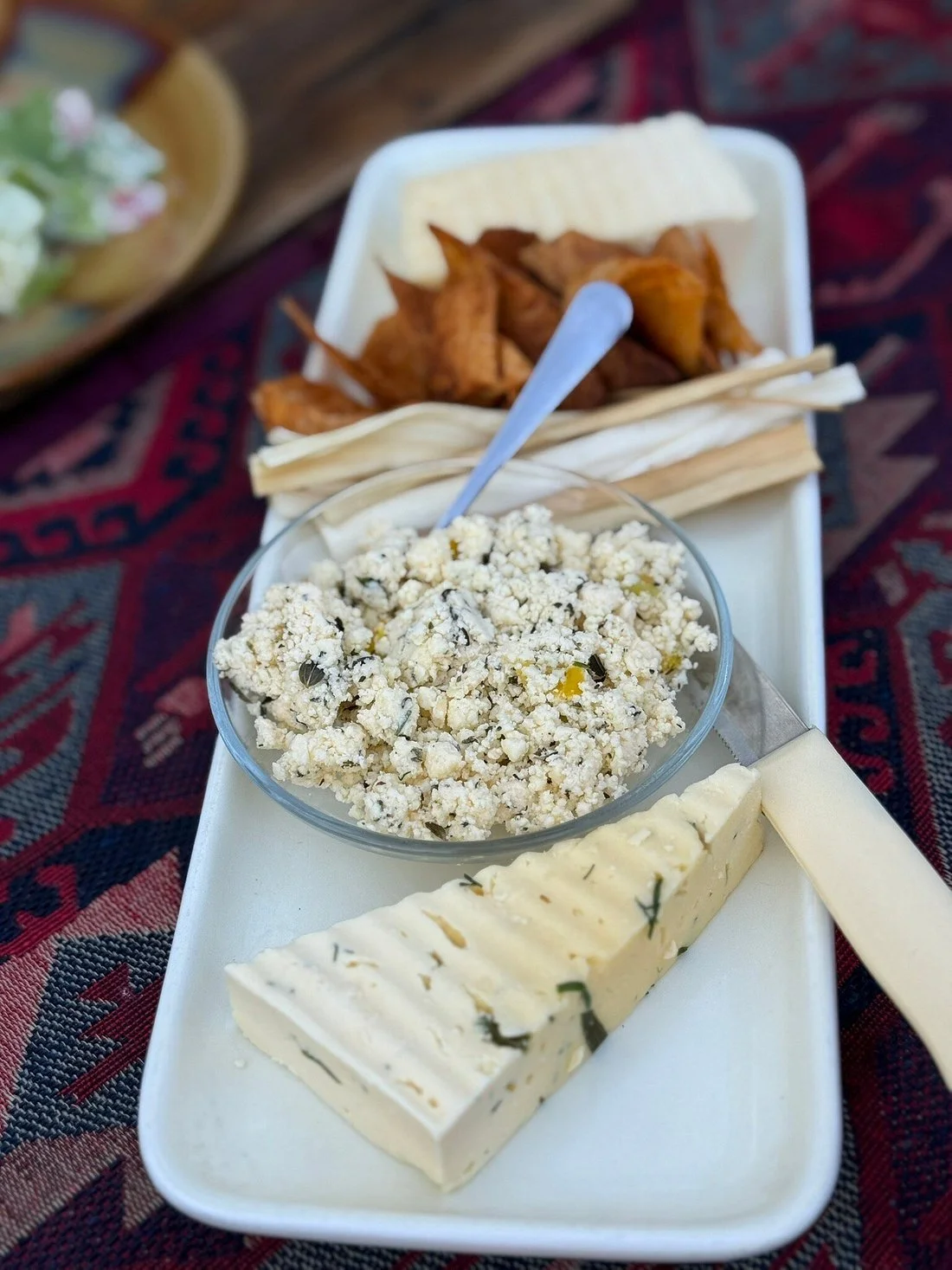 A plate of assorted cheeses, a bowl of crumbled blue cheese, some crackers, and fried items with a dip in the background, on a table with a patterned cloth.