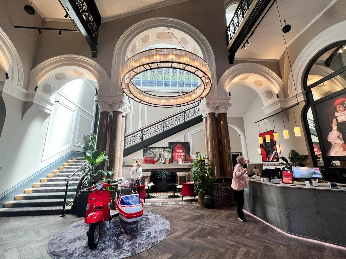 Lobby area with grand staircase, large chandelier, reception desk, and art decor. Children’s red ride-on motorcycle parked on a round rug, and staff member assisting at the counter.