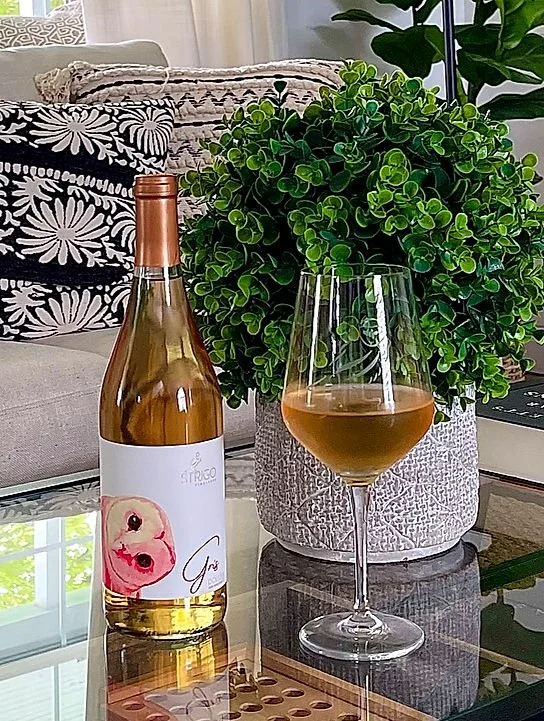 A bottle of rosé wine and a glass filled with rosé wine on a glass table. Behind them is a potted green bush and a cushioned sofa with black and white floral patterned pillows in a bright living room.