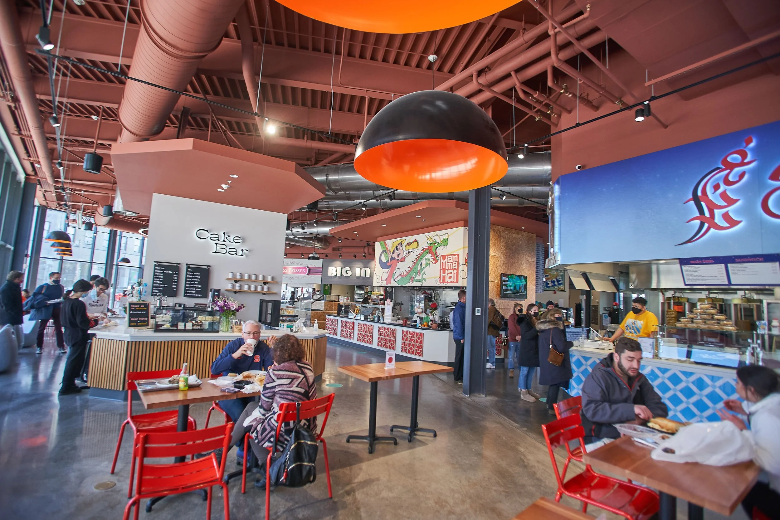 Indoor food court with customers dining and ordering at various counters, including a cake bar and Asian cuisine stall, under industrial ceiling with large black and orange pendant lights.