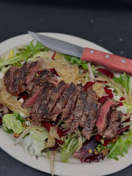 Steak slices on a bed of mixed salad greens with onions and cheese, with a knife resting on the plate.