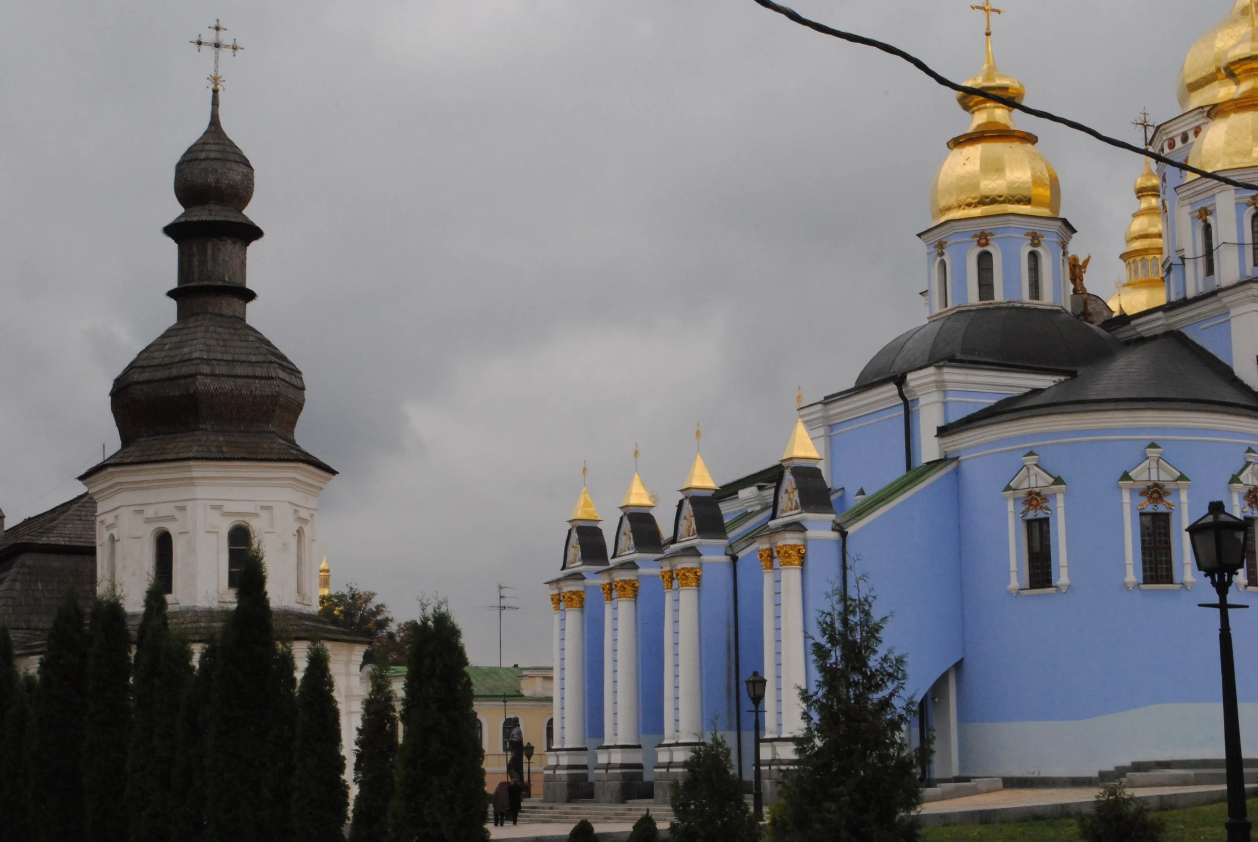 A church with a dark onion-shaped dome and a yellow and blue facade, topped with golden domes and crosses, under a cloudy sky.