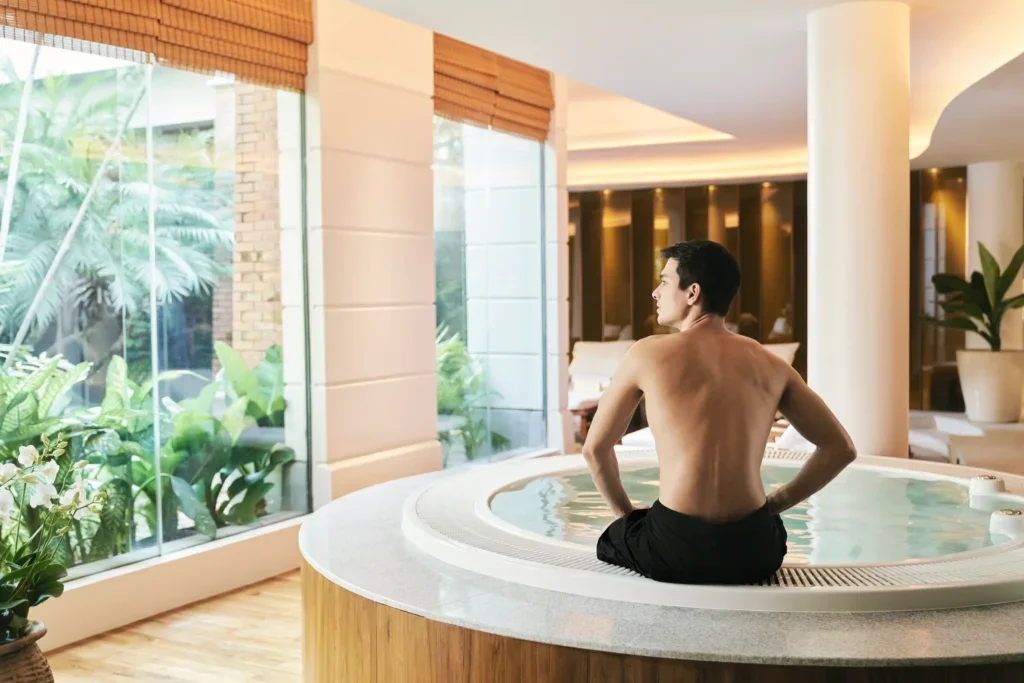 A man relaxing in a hot tub indoors with large windows showing greenery outside.