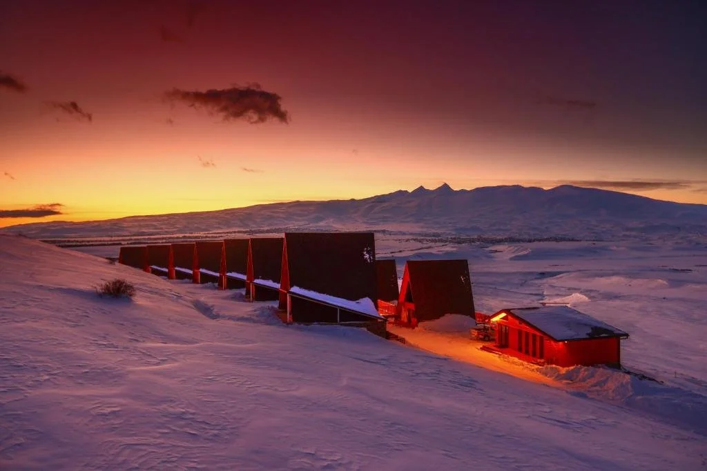 Snow-covered landscape featuring mountain scenery during sunset or sunrise with a line of dark red buildings, possibly a ski resort or cabin, lit with warm orange lights.