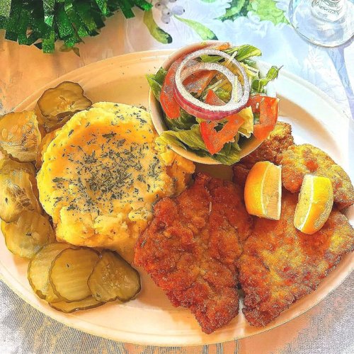A plate with fried fish fillets garnished with lemon wedges, a serving of mashed sweet potatoes sprinkled with herbs, sliced pickles, and a small bowl of mixed salad with onions, lettuce, and tomato. There are also two lemon wedges and a side of seasoned cucumbers.