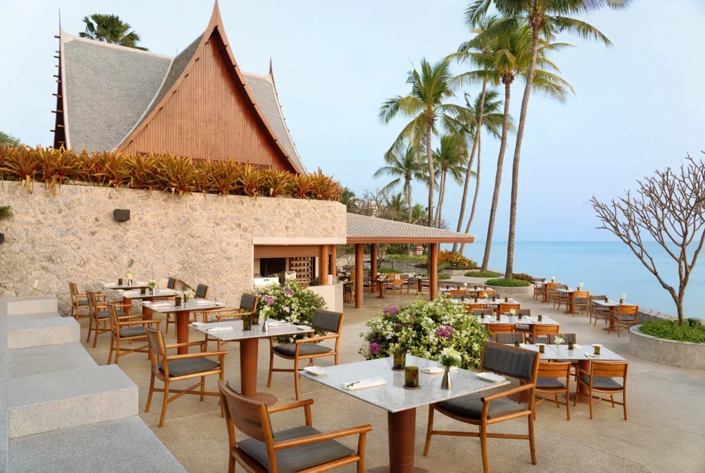 Outdoor restaurant with tables set for dining, overlooking the ocean, surrounded by palm trees and tropical plants, with a traditional wooden building in the background.