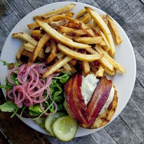 A plate of food with French fries, a salad with pickled onions and cucumber slices, and a grilled chicken breast topped with bacon and a fried egg.