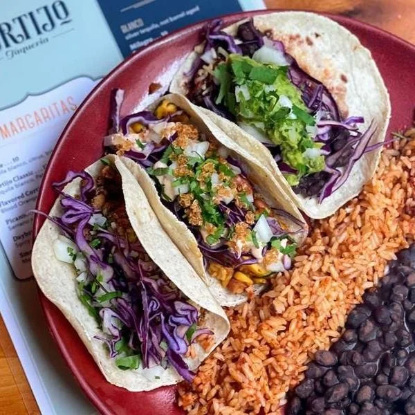 Three soft tacos filled with shredded lettuce, purple cabbage, corn, chopped onions, and fresh cilantro, served with Mexican rice and black beans on a red plate.