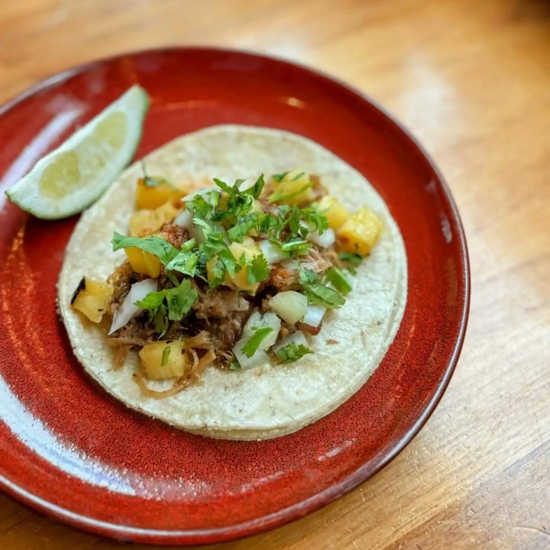 A taco on a red plate with chopped herbs and pineapple, accompanied by a lime wedge, on a wooden table.