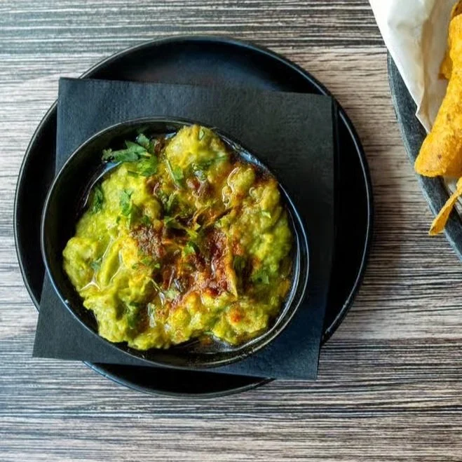 A bowl of guacamole topped with shredded cheese and cilantro, placed on a black circular plate with a napkin underneath, on a wooden table, with a side of chips visible to the right.