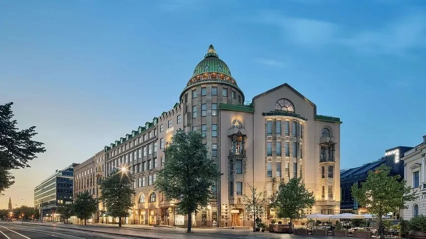 A historic building with a green dome and rounded tower, illuminated at dusk, on a city street lined with trees and modern buildings.