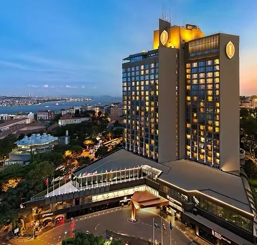 A tall hotel building with illuminated windows and the CoinMarketCap logo on top, located in a cityscape during dusk with a view of a bay and other buildings in the background.