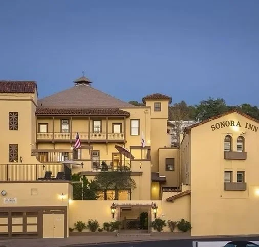 Exterior of the Sonora Inn, a yellow building with multiple levels and a Spanish-style terracotta roof, with balconies, flags, and a gated entrance.