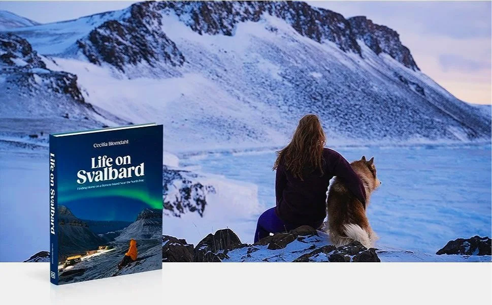 A woman with long hair sitting on rocks next to a dog, overlooking a snowy mountainous landscape with glaciers.