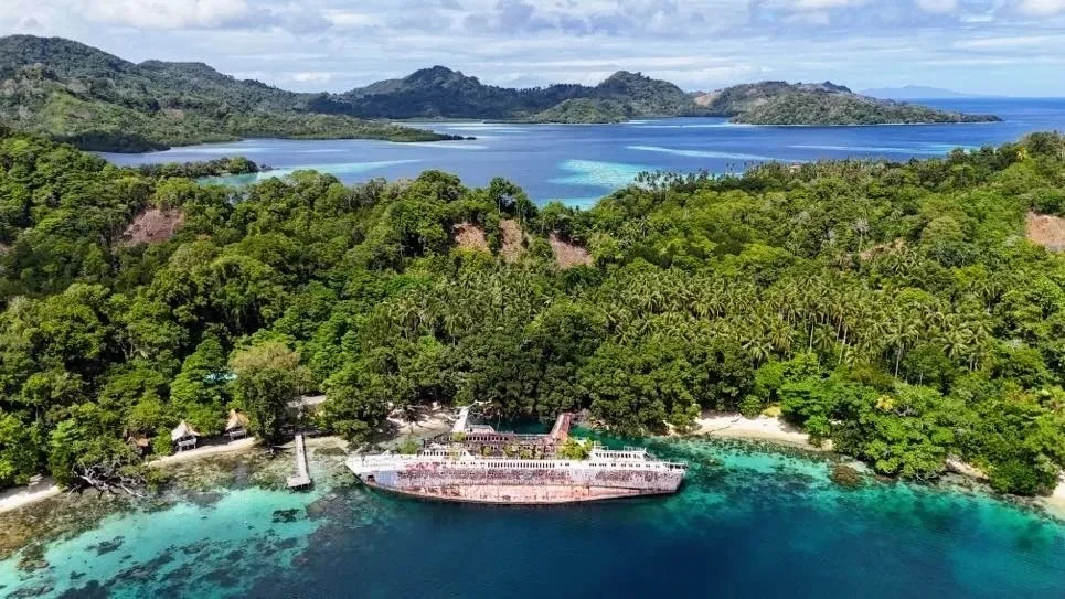 A lush green island with a shipwreck on a sandy beach in clear blue waters, surrounded by other islands and a calm ocean.