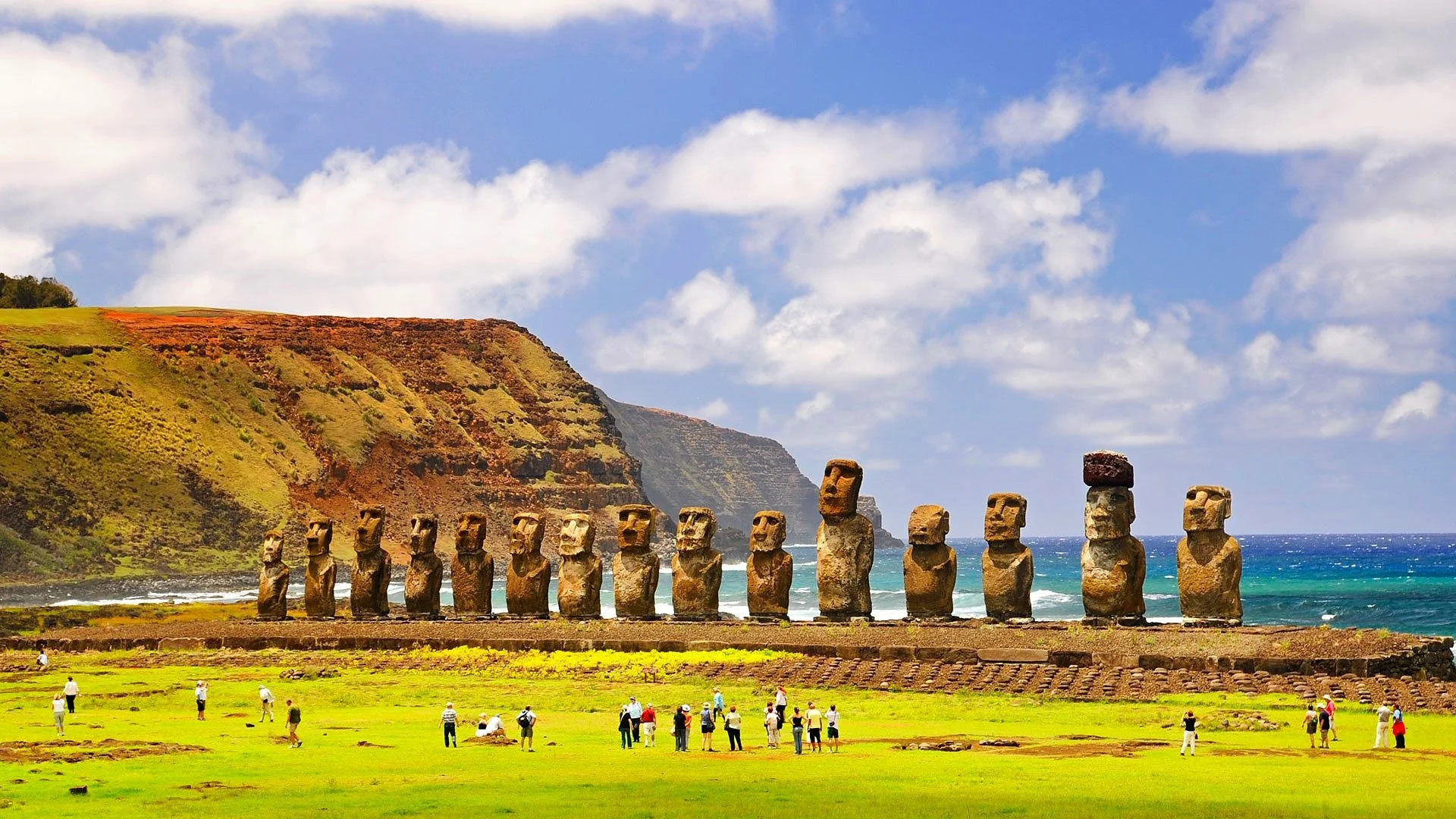 Moai statues on Easter Island with tourists walking on green grass, coastal cliffs, blue ocean, and partly cloudy sky in the background.