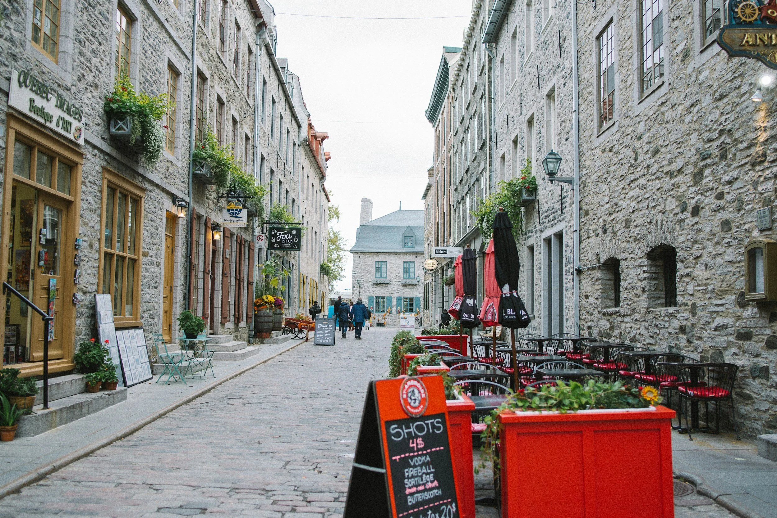A cobblestone street in a charming historic neighborhood with outdoor cafe seating on the right, featuring red chairs and black umbrellas. The street is lined with stone buildings with flower boxes and signs for shops and cafes, with a few people walking in the distance.