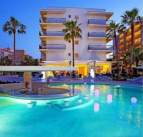 Modern hotel with a swimming pool surrounded by palm trees and outdoor seating, with a multi-story building in the background during evening hours.