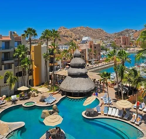 Resort pool area with a thatched roof, palm trees, lounge chairs, umbrellas, and a mountain backdrop under a clear blue sky.