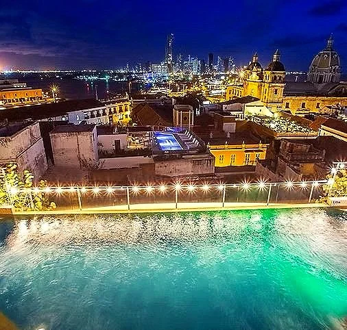 Nighttime cityscape with buildings, a rooftop pool lit with underwater lights, and a skyline with tall skyscrapers in the distance.