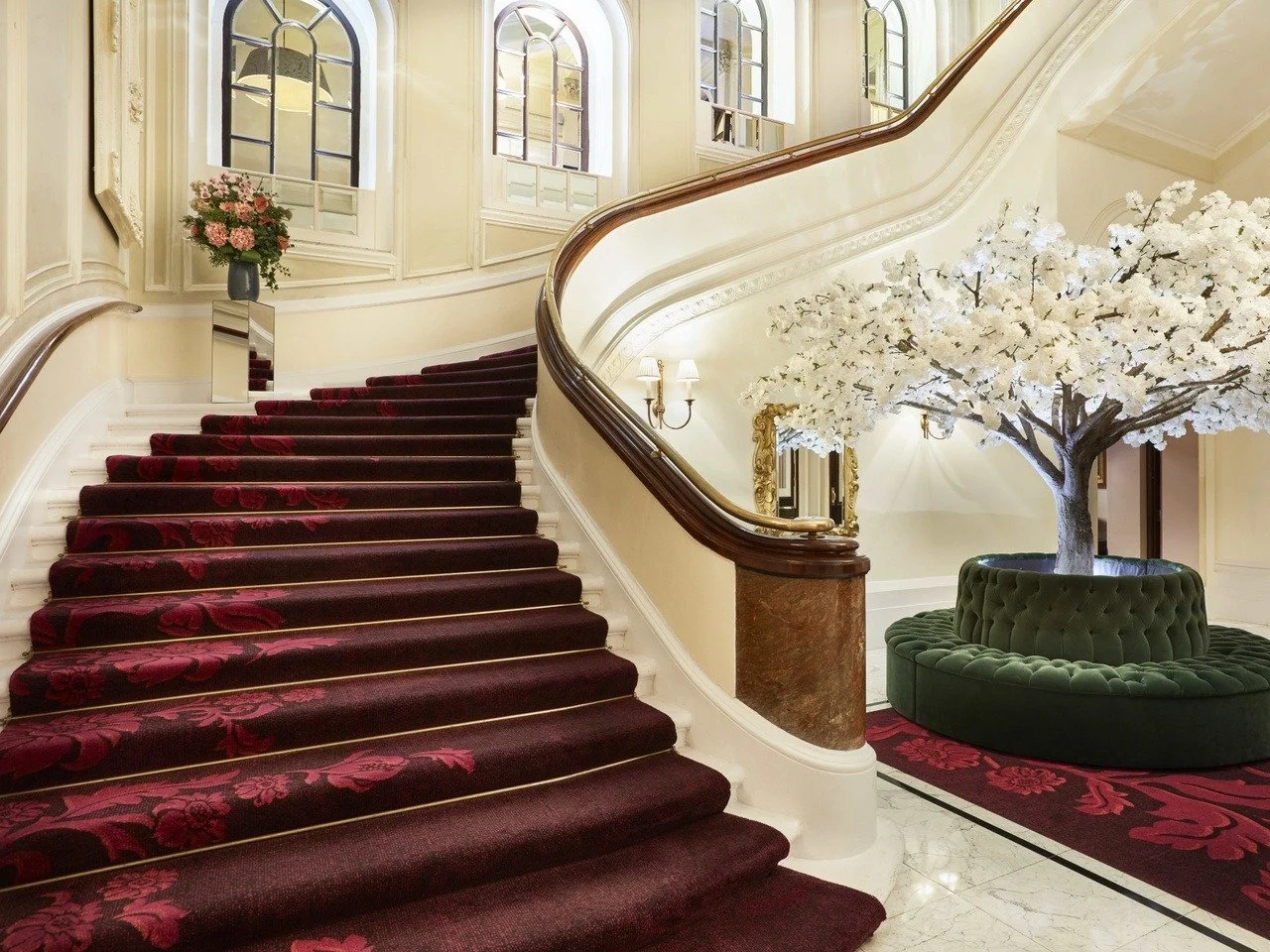 Elegant hotel lobby with a grand staircase with red carpet, white walls, large windows, a flower arrangement, and a decorative tree with white blossoms in a green velvet circular seating area.