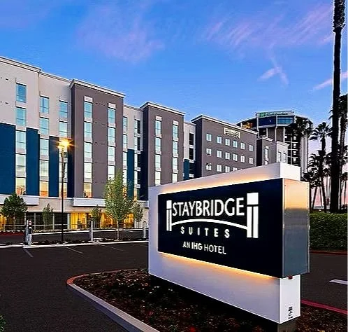 Exterior of Staybridge Suites hotel with a large sign in the foreground and a modern building in the background during sunset.