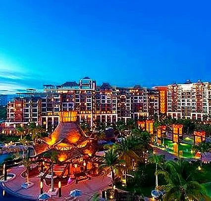 Night view of a hotel resort with illuminated pools, palm trees, and buildings under a dark blue sky.