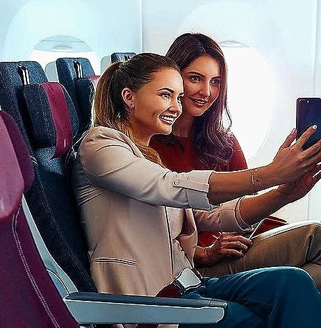 Two women sitting next to each other on an airplane, taking a selfie with a smartphone, smiling.