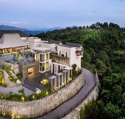 Luxury modern house on a hillside with a winding road, surrounded by lush green trees and mountains in the background.
