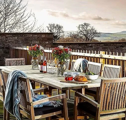 Outdoor dining table set on a patio with six wooden chairs, a floral centerpiece, a loaf of bread, tomatoes, wine bottles, and a jacket draped over one chair, with a wooden fence and trees in the background.