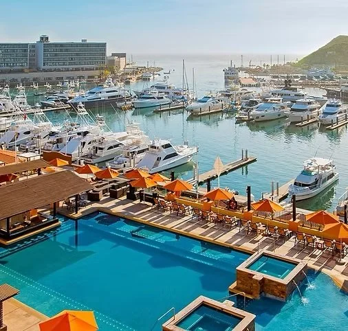 View of a marina with boats docked, a swimming pool with orange umbrellas and seating area, and a cityscape in the background.