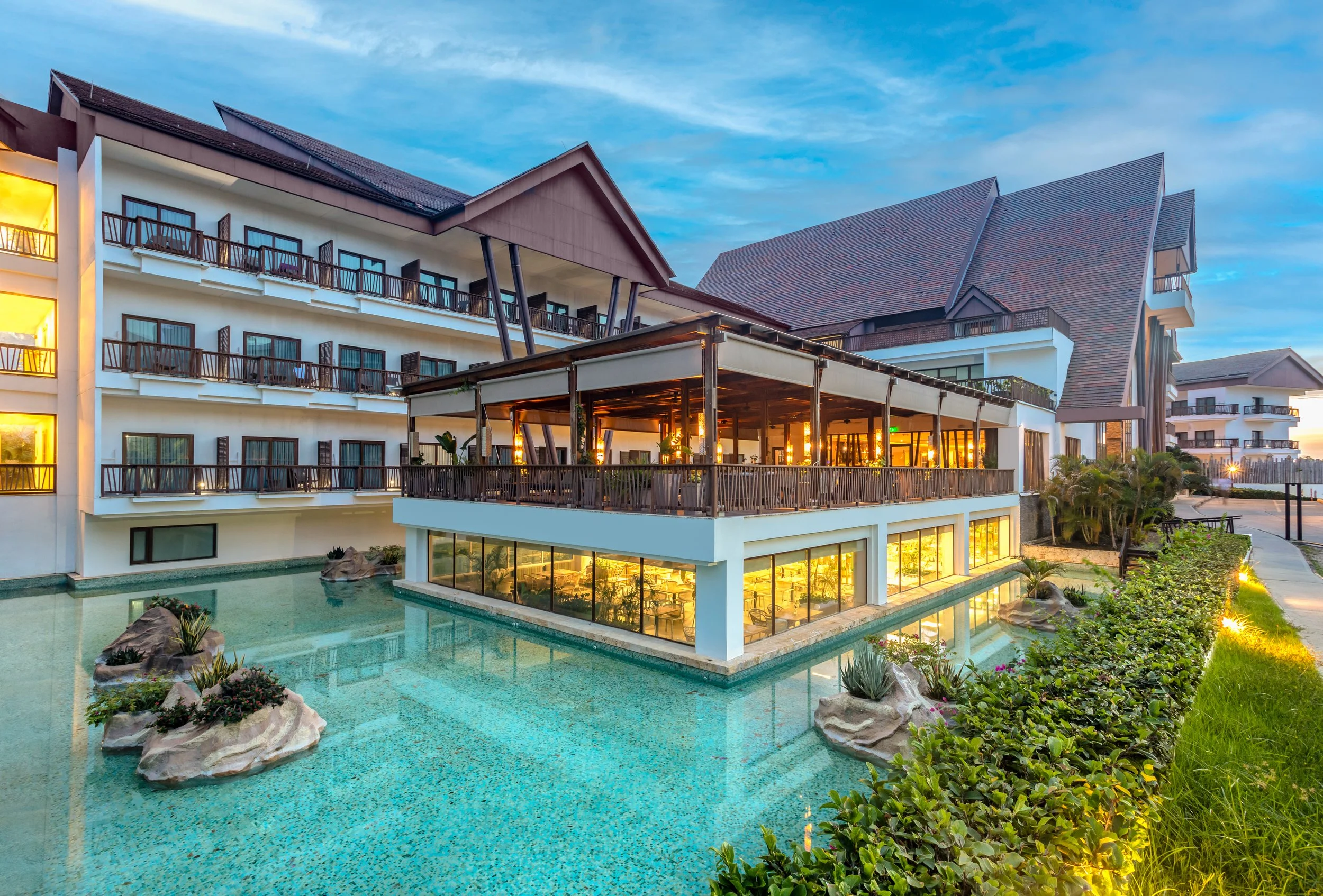 A modern hotel with multiple floors, balconies, and a large outdoor pool surrounded by rocks and greenery, illuminated by warm lighting at dusk.