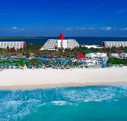 A beachfront resort with white sand beach, turquoise water, and multiple white buildings with red accents and a central red-roofed structure, under a blue sky.