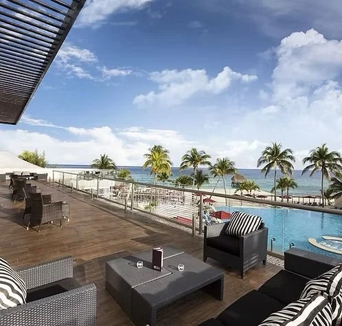 Poolside lounge area with black wicker chairs and tables overlooking a beach with palm trees and ocean, under a partly cloudy sky.